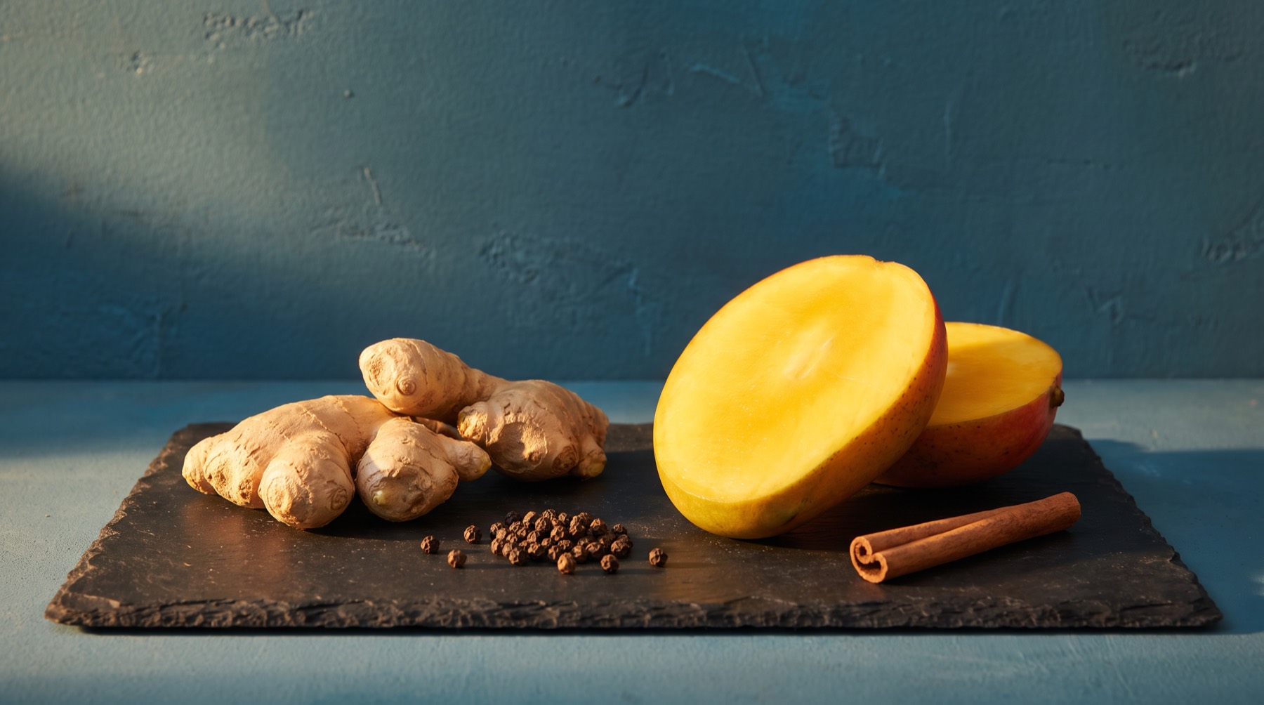 Fresh ginger root, black peppercorns, a cinnamon stick and a halved mango on a dark slate board
