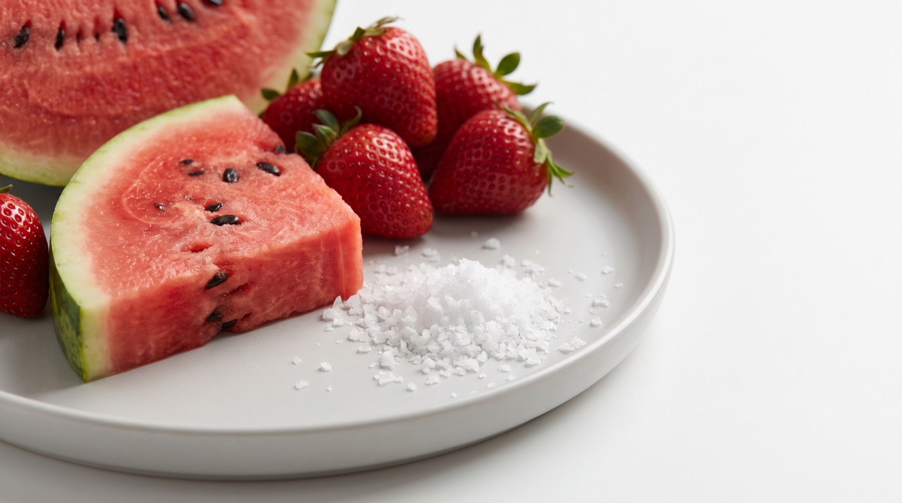 Watermelon wedge and strawberries on a grey plate beside a small mound of flaky sea salt