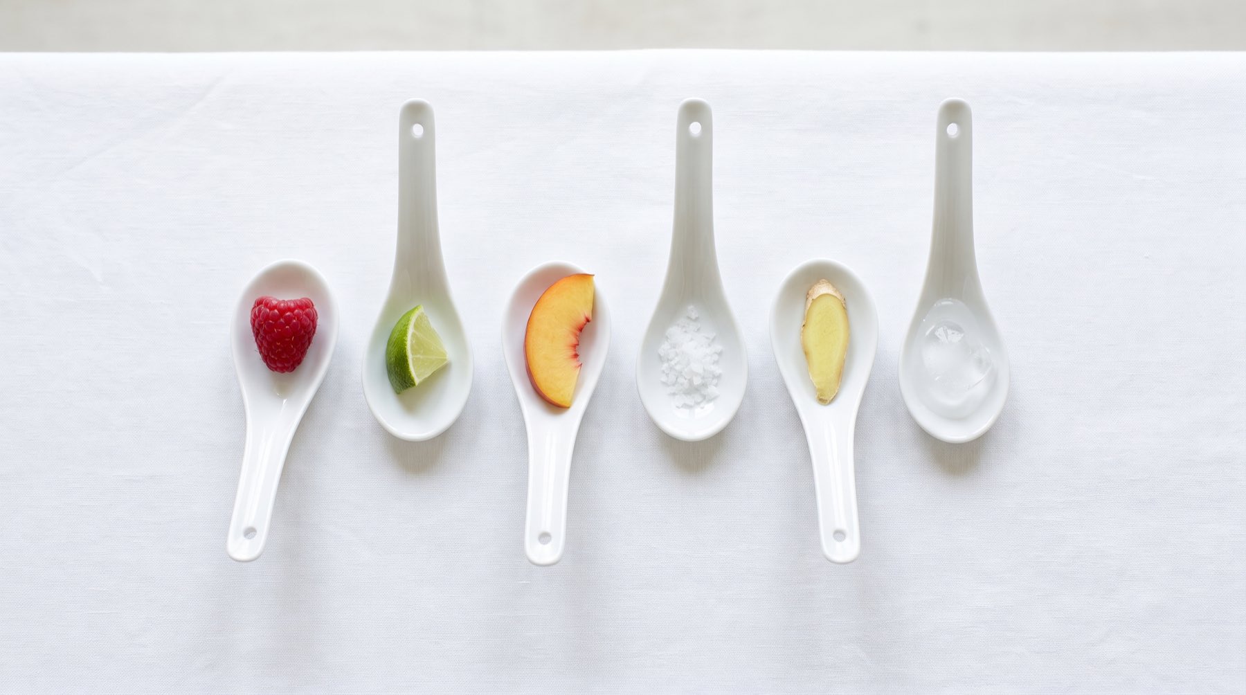 A row of small white tasting spoons on white linen, each holding a raspberry, a lime segment, a slice of peach, salt flakes, ginger or an ice chip